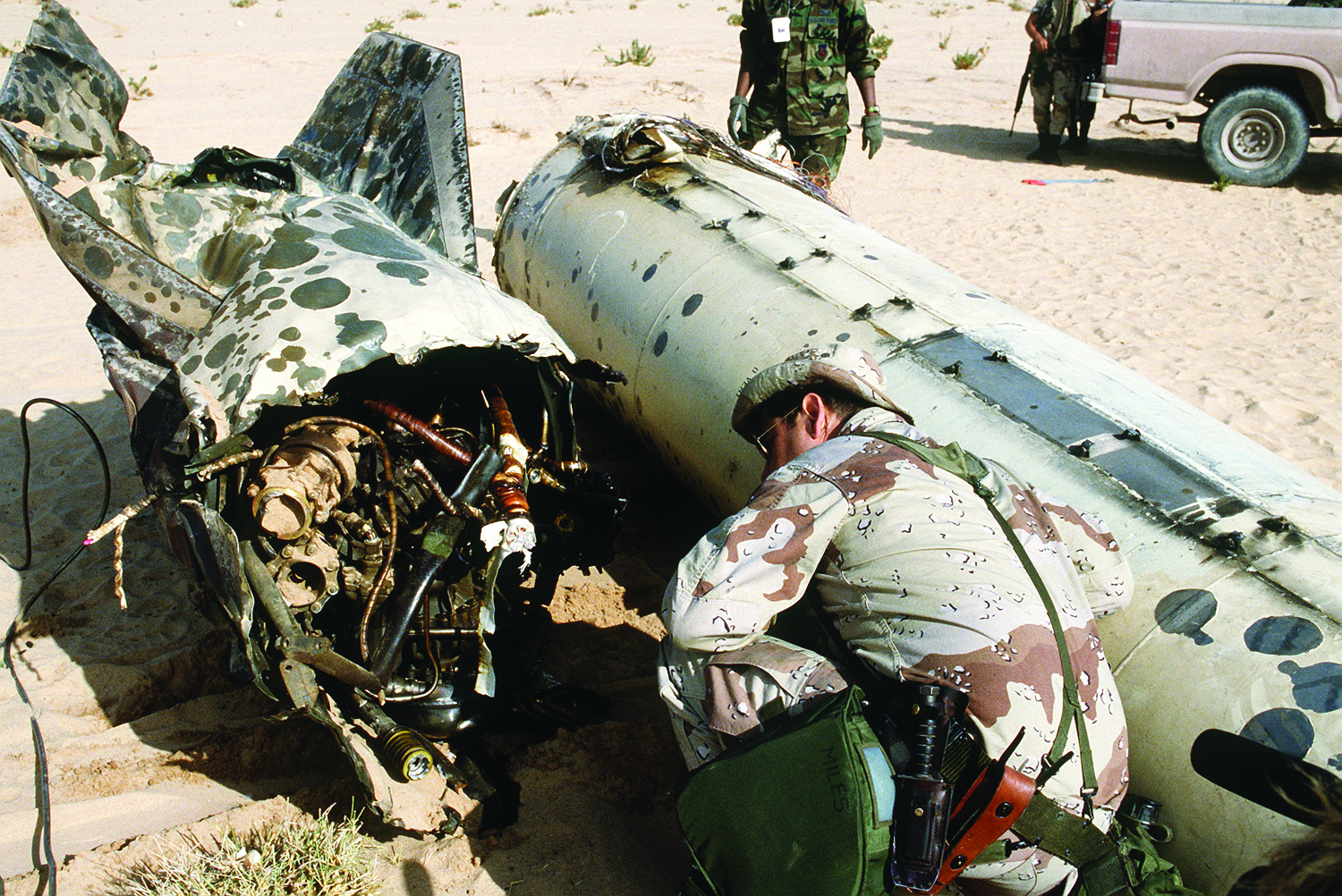 A soldier in desert camouflage inspects the wreckage of a Scud missile that has been destroyed. The missile is broken in half on the sand, exposing its internal wiring and components.